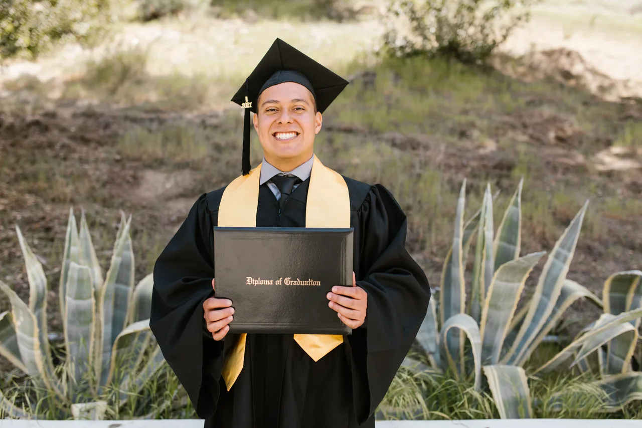 A young man in graduation attire happily holding his diploma outdoors