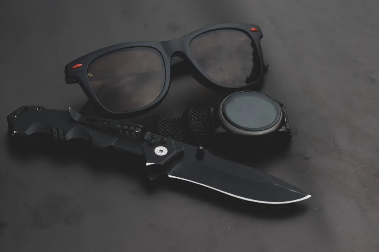 A minimalist black still life featuring sunglasses, a knife, and a wristwatch on a dark surface