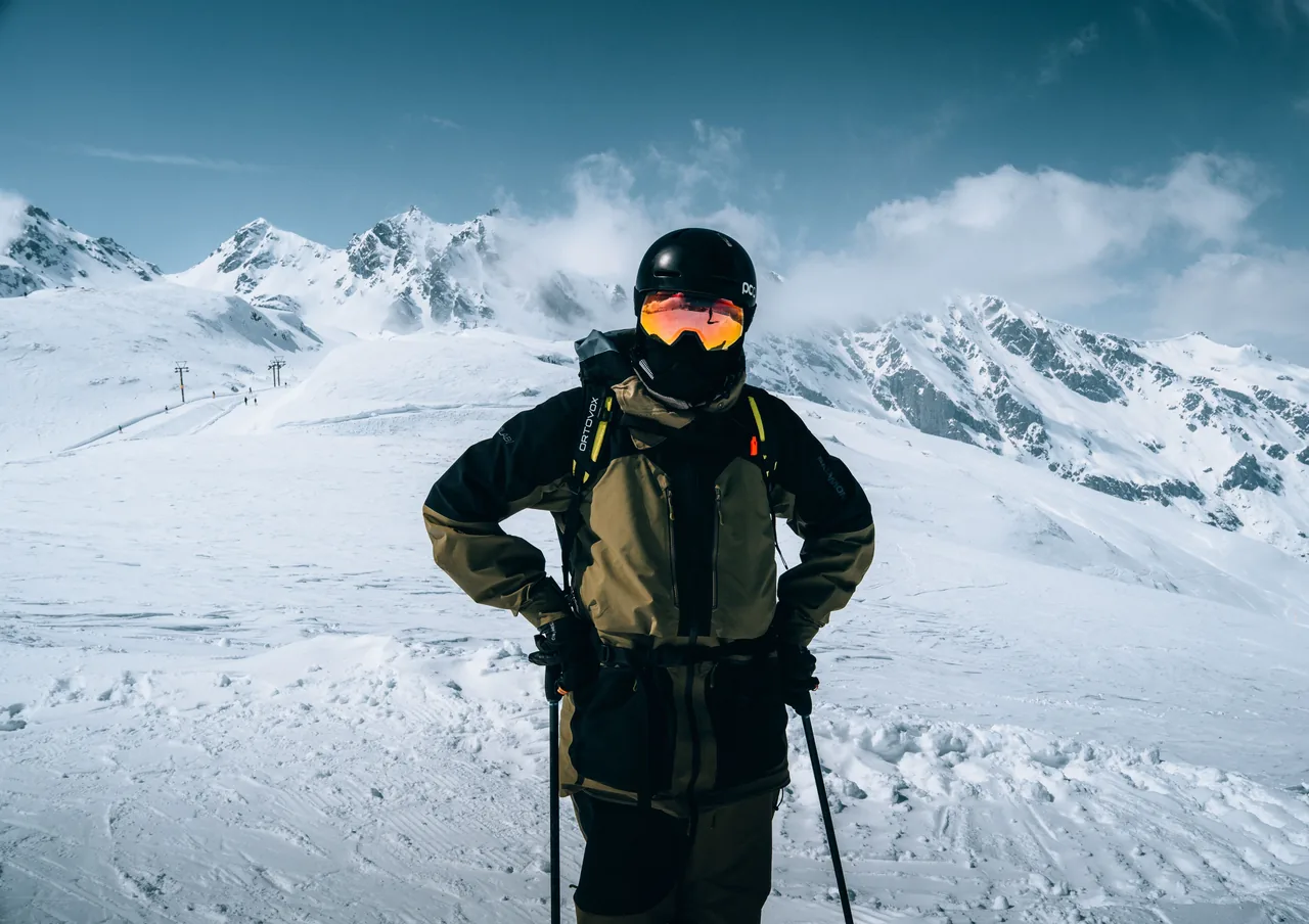 A skier stands confidently in full gear against a backdrop of snow-covered alpine peaks
