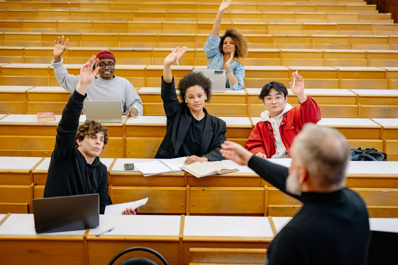 A diverse group of university students participating in a lecture, raising hands to engage with the professor