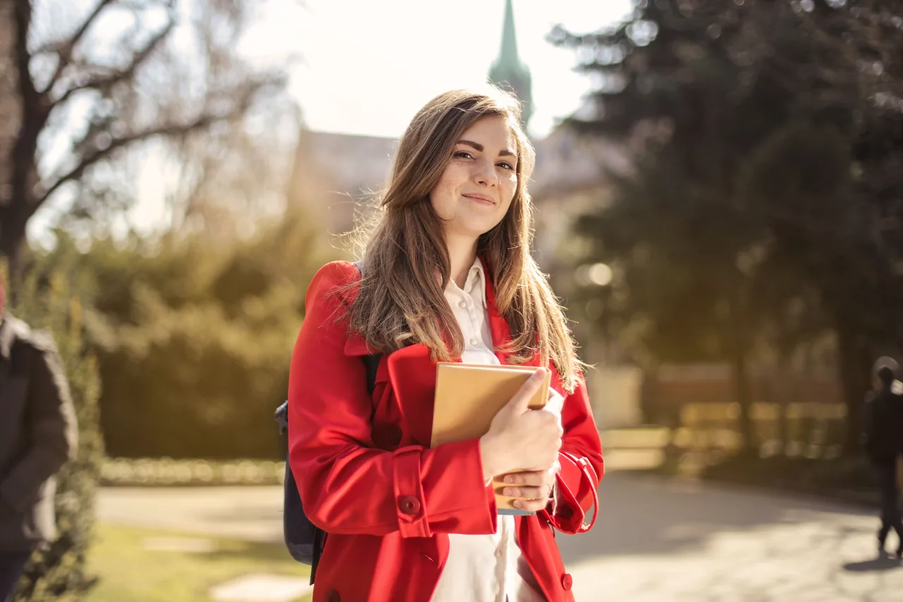 Young woman in a red coat smiling while holding books outdoors on a sunny day