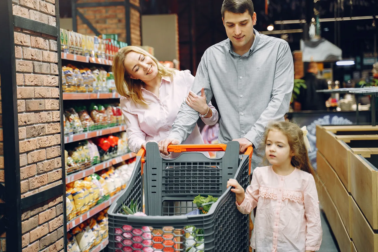 A family of three shopping in a supermarket, pushing a cart full of groceries